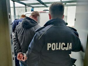 Dwóch umundurowanych policjantów odwróconych tyłem prowadzi do radiowozu zatrzymanego mężczyznę, który stoi pośrodku policjantów.