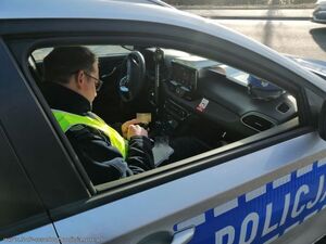 Policjant siedzi w radiowozie oznakowanym, ma opuszczoną szybę.