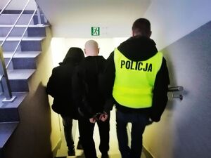 Dwóch policjantów schodzi po schodach z zatrzymanym mężczyzną, który idzie w środku. Policjant z prawej strony ma założoną żółtą kamizelkę z napisem policja.