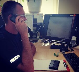 Policjant siedzi przy biurku na którym ustawione są monitory komputerów i trzyma słuchawkę od telefonu przy uchu.
