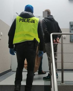 dwóch policjantów wchodzi z zatrzymanym mężczyzną po schodach na górę, zatrzymany trzyma ręce w kajdankach z tyłu