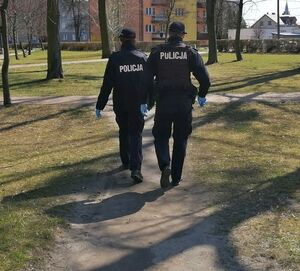 Umundurowani policjanci idą ścieżką w parku, są odwróceni tyłem.