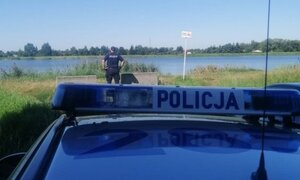 Policjant stoi na brzegu zbiornika i patrzy przed siebie, stoi odwrócony tyłem, za nim stoi zaparkowany radiowóz.