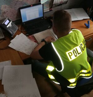 Policjant w żółtej kamizelce z napisem policja siedzi przy biurku przed komputerem.