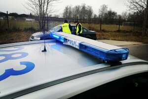Dwóch umundurowanych policjantów stoi przy zaparkowanym samochodzie, przed nimi widoczny sygnalizator z napisem policja na dachu radiowozu.