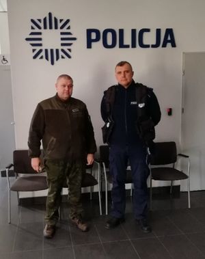 Umundurowany policjant stoi po prawej stronie, obok niego stoi umundurowany strażnik łowiecki, nad funkcjonariuszami na ścianie znajduje się napis policja.