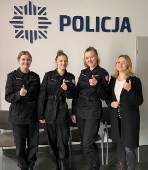 Uczennice z pracownicą komendy po prawej stronie stoją przy ścianie z napisem Policja.