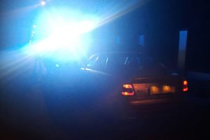 miejsce interwencji na drodze, srebrny samochód stoi na ulicy, obok niego migają policyjne niebieskie światła