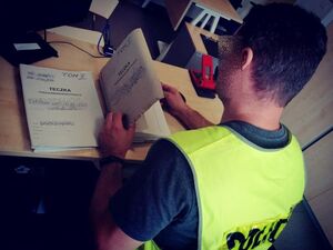 Policjant odwrócony tyłem w żółtej kamizelce siedzi przy biurku i przegląda akta.