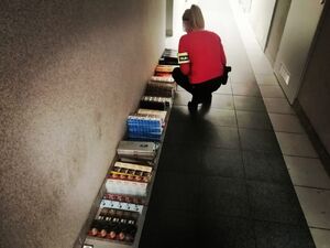 Policjantka z opaską na ramieniu z napisem policja pochylona nad papierosami nielegalnymi ułożonymi pod ścianą na korytarzu.