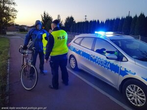 Policjant stoi przy radiowozie odwrócony do zdjęcia tyłem i podaje kamizelkę odblaskową rowerzyście stojącym przed nim