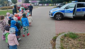 dzieci ustawione w parach stoją obok zaparkowanego radiowozu a przed nimi stoi policjantka