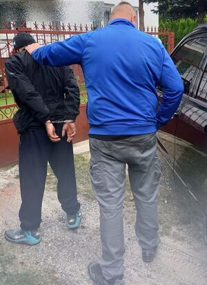 policjant w niebieskiej bluzie stoi odwrócony tyłem i trzyma zatrzymanego mężczyznę stojącego przed nim, który ma ręce spięte z tyłu