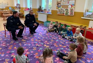 policjanci siedzą na małych krzesełkach ustawionych na środku sali a przed nimi siedzą w grupie małe dzieci