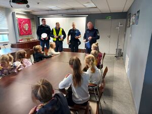 policjanci stoją na środku sami a komendant podchodzi do siedzących przy stolikach dzieci i rozdaje upominki