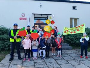 Dwóch policjantów w żółtych kamizelkach stoi wraz z dziećmi i nauczycielami z transparentami przedstawiającymi jabłko i cytrynę