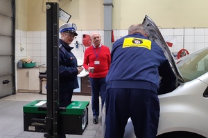 Mężczyzna odwrócony tyłem do zdjęcia ustawia światła pod otwartą maską samochodu, obok stoi policjantka w mundurze i białej czapce oraz drugi mężczyzna w czerwonej bluzie