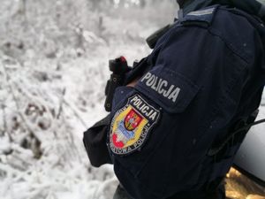 Ramię umundurowanego policjanta z naszywką na mundurze. Policjant stoi przed zaparkowanym radiowozem, widoczne drzewa pokryte śniegiem