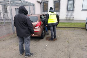 Dwóch policjantów stoi obok brązowego samochodu przy otwartych tylnych drzwiach