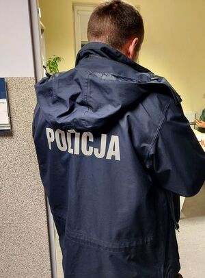 Policjant odwrócony tyłem stoi w drzwiach, na plecach kurtki ma biały napis policja,