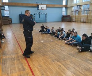 policjant stoi na środku sali gimnastycznej i mówi do dzieci siedzących przed nim na podłodze
