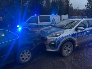 Samochód koloru ciemnego zatrzymany po pościgu, stoi uszkodzony, obok niego radiowozy policyjne z migającymi niebieskimi światłami.