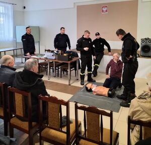 Policjanci i strażacy stoją na środku sali, w której na krzesełkach siedzą zgromadzeni mieszkańcy.