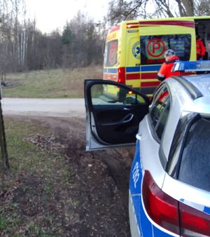 Radiowóz policyjny z otwartymi drzwiami od strony kierowcy stoi tuż za żółtą karetką pogotowia przy której stoi medyk w pomarańczowym kombinezonie.