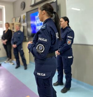 Trzy policjantki i jedna kobieta stoją na środku klasy w której przed nimi siedzą w ławkach uczniowie klas mundurowych, uczniowie są w czarnych mundurach.