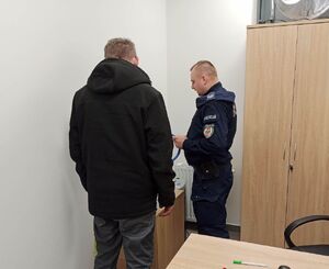 Policjant w granatowym mundurze stoi ustawiony bokiem obok niego stoi odwrócony tyłem mężczyzna w czarnej bluzie.