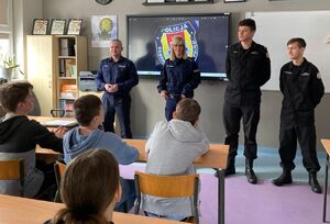 Policjant i policjantka stoją na środku klasy, w której przy ławkach siedzi młodzież.