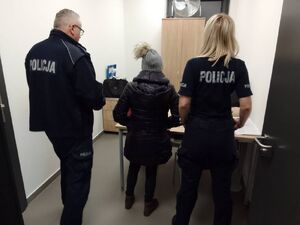 Zatrzymana kobieta stoi w środku pomieszczenia obok niej po prawej stronie policjantka a po lewej policjant, wszyscy są odwróceni tyłem.