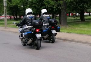 dwóch motocyklistów jedzie drogą obok siebie w czarnych skórzanych kurtkach z napisem policja i białych kaskach