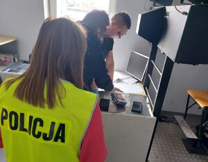 Kobieta w żółtej kamizelce z napisem policja stoi w pomieszczeniu z dwoma mężczyznami gdzie jeden pobiera od drugiego odciski palców.