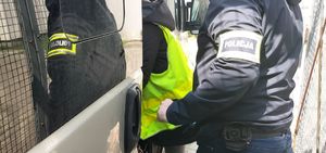 Policjant w żółtej kamizelce z napisem policja prowadzi zatrzymanego mężczyznę do szarego samochodu.