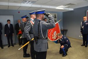 Uroczysta zbiórka na sali odpraw stoją umundurowani policjanci, stoi poczet sztandarowy i zaproszeni goście, jeden z policjantów klęka schylony do pochylonego sztandaru.