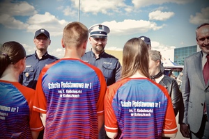Grupa trzech osób młodych stoi odwrócona tyłem, na ich koszulkach na plecach widnieje napis Szkoła Podstawowa numer 1 w Koluszkach, młodzi ludzie stoją przed umundurowanymi policjantami.