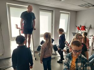 Grupa dzieci jest w pomieszczeniu siłowni, w którym ustawione są sprzęty do ćwiczeń, obok dzieci stoją umundurowani policjanci i mężczyzna w stroju sportowym.