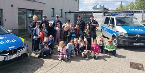Dzieci z kobietą stoją w grupie przed budynkiem obok którego stoją zaparkowane radiowozy policyjne a za dziećmi stoi czterech policjantów i jedna policjantka.