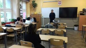 Policjantka stoi na środku klasy przed nią siedzą osoby dorosłe w ławkach klasowych.