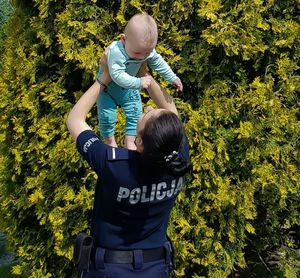 Policjantka w granatowym mundurze podnosi rękoma do góry małego niemowlaka.