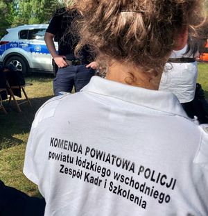 Plecy odwróconej kobiety, która ma białą koszulkę z napisem Komenda Powiatowa Policji Powiatu Łódzkiego Wschodniego Zespół Kadr i szkolenia.