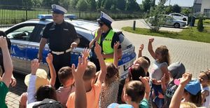 Policjant i policjantka w białych czapkach stoją przed dziećmi które podnoszą ręce do góry, za policjantami ustawiony jest radiowóz.