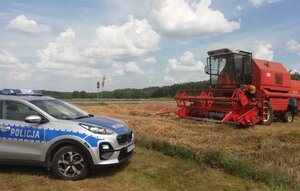 Radiowóz stojący na polu zboża a przed nim czerwony duży kombajn.