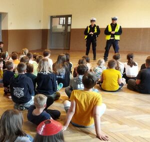 Policjant i policjantka z białymi czapkami na głowie stoją na środku sali, w której siedzą przed nimi dzieci.
