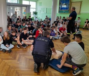 Policjant kuca przy chłopcach i leżącym na podłodze fantomie, przed nim siedzi duża grupa dzieci.