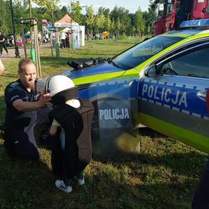 Policjant kuca przy małych chłopcu, któremu zakłada policyjną kamizelkę i biały kask na głowę, obok stoi radiowóz i wóz strażacki.