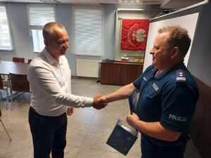 Policjant w granatowym mundurze podaje rękę mężczyźnie w białej koszuli, który stoi na przeciwko niego.