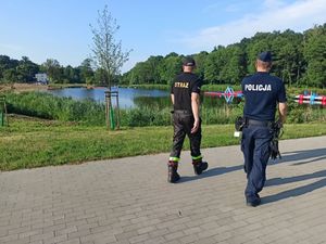 Policjant i strażak idą chodnikiem wzdłuż zbiornika wodnego i patrzą w jego stronę.