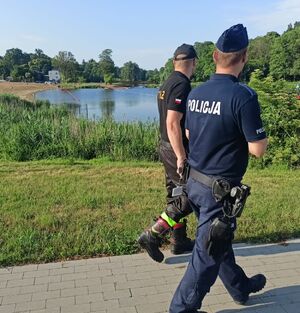 Policjant i strażak idą chodnikiem wzdłuż zbiornika wodnego i patrzą w jego stronę.